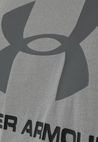 Camiseta gris de manga corta con un gran logo oscuro y el nombre de la marca impresos en la parte inferior, hecha de una tela suave y ligera.