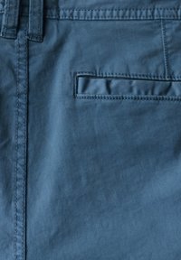 Modrá bavlněná látka zblízka ukazuje hladkou strukturu, detaily švů a malou obdélníkovou kapsu. Látka má mírný lesk.