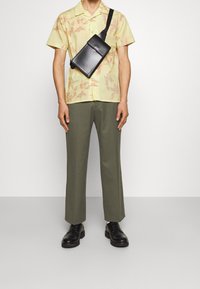 Korte mouwen, lichtgele shirt met een bloemenpatroon; olijfgroene op maat gemaakte broek; zwarte schoudertas; zwarte veterschoenen.