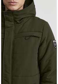 Olivenfarget boblejakke med høy krage, glidelås og brystlomme med logo-patch. Strukturert stoff med horisontale sømmer.