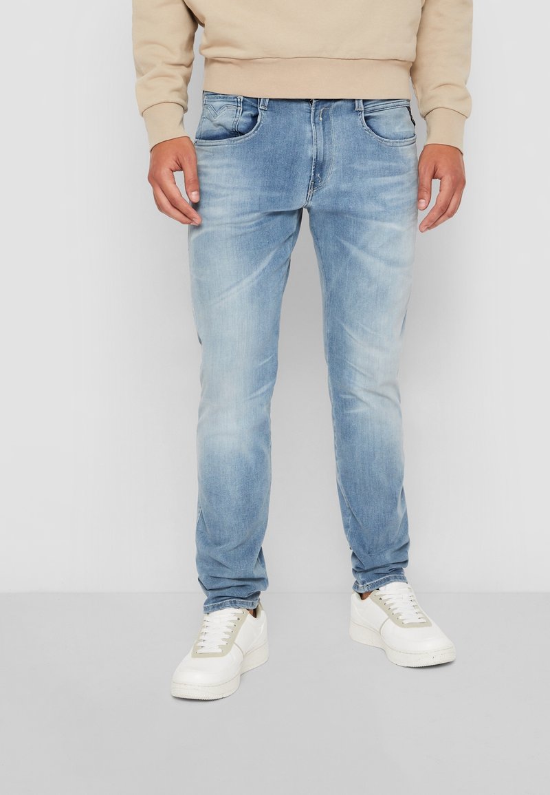 Jean en denim bleu clair avec une coupe slim, doté de poches avant et de détails délavés. Associé à des baskets blanches et un sweat beige.