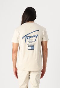 Krémové bavlněné tričko s velkým modrým potiskem s nápisem "Tommy" a názvy měst. Krátké rukávy a volný střih. Spárováno se světlými kalhotami.