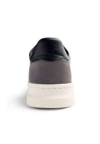 Zapatilla de ante gris con detalles en piel negra, talón redondeado y suela blanca texturizada. Presenta un logo sutil en relieve en el talón.