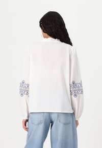 Blouse blanche avec un col à volants et des manches longues, ornée de motifs floraux bleus brodés sur les manches. Tissu doux, coupe décontractée.