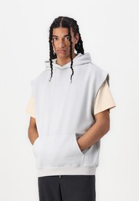 Hellgrauer ärmelloser Hoodie mit einer Fronttasche, aus einem Baumwoll-Mischgewebe, mit weicher Textur und großer Kapuze, getragen über einem beigen Kurzarmshirt.
