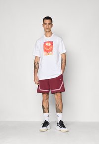 Witte T-shirt met kleurrijke grafische print, gecombineerd met bordeauxrode sportshorts met contrasterende rand en zwarte sneakers met witte zolen.