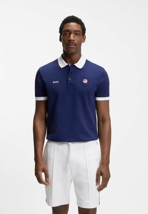 Man staat, draagt een donkerblauw poloshirt met witte kraag en een Amerikaan vlag patch, gecombineerd met witte short met trekkoord.