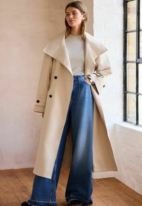 Beige trenchcoat med brede klapper og dobbeltradet design. Bæres med højtsiddende blå vidde bukser. Glat tekstur med knapdetaljer.