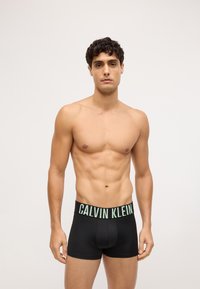Crne bokserice od glatkog materijala s širokim zelenim pojasom na kojem piše "CALVIN KLEIN". Model stoji s neutralnim izrazom lica.