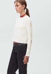 Ženska v beli dolgem rokavu majici s odprtinami na ramah, črnih kavbojkah s kovinskimi nitmi, rdeči choker verižici in rdeči zapestnici, stoji v profilu.