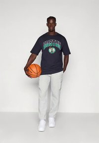 Μοντέλο που φοράει μια ναυτική μπλουζάκι oversized με κείμενο και γραφικό "BOSTON", συνδυασμένο με ανοιχτοί γκρι cargo παντελόνια φόρμας και λευκά αθλητικά παπούτσια, κρατώντας μια μπάλα μπάσκετ.