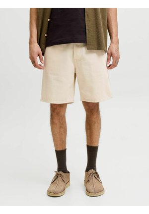 Férfi, bézs térdig érő rövidnadrágban, sötét zokniban és világosbarna fűzős cipőben, áll egy egyszínű világos háttér előtt.