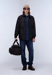 Svart jacka med flera fickor över en jeansskjorta, blå jeans och bruna skor. Modellen håller en svart duffelväska, bär solglasögon och en beige mössa.