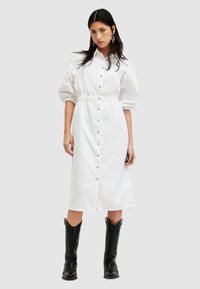 Weißes Midi-Kleid mit Knopfverschluss, Puffärmeln und elastischem Bund. Kombiniert mit schwarzen kniehohen Cowboy-Stiefeln.