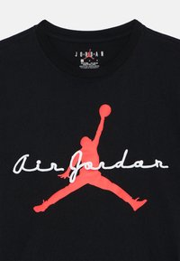 Μαύρο βαμβακερό μπλουζάκι με κόκκινο λογότυπο Jumpman και λευκό κείμενο "Air Jordan". Στρογγυλή λαιμόκοψη, κοντά μανίκια, λεία υφή.