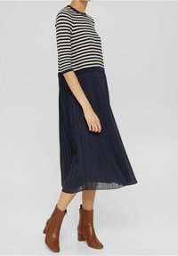 Ränderad marinblå och krämfärgad klänning med en plisserad marinblå kjol; har en åtsittande topp och textilmaterial, ihop med bruna ankelstövlar.