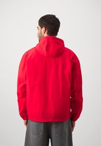 Röd lättviktsjacka med huva, ripstop-struktur, elastiska ärmslut och minimalistisk design. Kombinerad med grå löst sittande shorts.