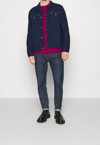 Giacca in denim blu scuro con due tasche sul petto, abbinata a un maglione magenta sotto e jeans scuri, completata da scarpe nere con lacci.
