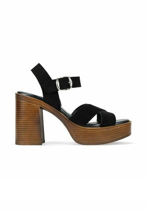 Sándala de tacón alto de ante negro con un tacón de bloque de madera, que presenta una correa con una hebilla de plata y correas frontales cruzadas.