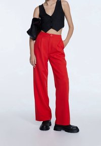 Osoba oblékající černý pruhovaný bezrukávový crop top, červené kalhoty s vysokým pasem a širokými nohavicemi, černé masivní boty a držící černou tašku pod jednou paží.