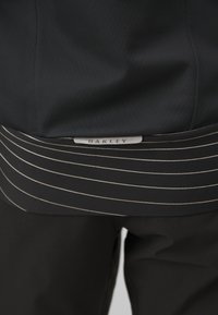Juodas švarkas su tekstūruota apdaila. Yra dryžuotas audinio apvadas ir "OAKLEY" etiketė. Lygus medžiaga su prigludusia forma.