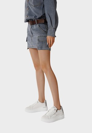 Persona con gonna e giacca in denim azzurro chiaro con cintura marrone, abbinati a sneakers bianche con plateau, in piedi davanti a uno sfondo semplice.