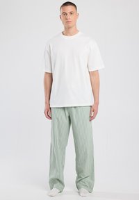 Man staat rechtop met de voorkant naar voren, gekleed in een eenvoudig wit oversized T-shirt, lichtgroene en witte verticaal gestreepte losse broek, en witte sokken.