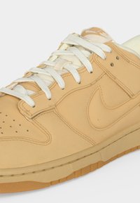 Χαμηλά ταμπά αθλητικά παπούτσια Nike με λευκά κορδόνια, κεντημένο λογότυπο Swoosh στο πλάι και σόλα από καουτσούκ σε λευκό φόντο.