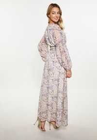 Lange, met bloemen bedrukte jurk in pasteltinten, met lange mouwen, een taille die is aangesnoerd en een vloeiende silhouette; gedragen met gouden hoge hakken.