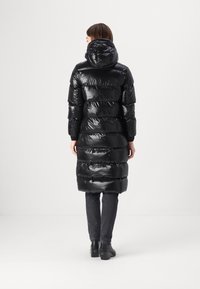 Lang zwarte pufferjas met een glanzende afwerking, voorzien van een capuchon, horizontaal gestikte delen en elastische manchetten, gecombineerd met donkere jeans en enkellaarsjes.