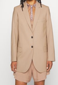 Blazer marron clair à deux boutons, poches latérales et revers nets, porté sur une chemise beige scintillante et associé à un short ajusté.