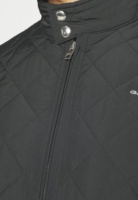 Black quilted jacket featuring a high collar, silver snap closures, and a central zipper. Smooth texture with a modern design.