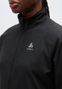 Gros plan sur une personne portant une veste Odlo noire zippée avec un col montant, montrant le logo de la marque sur la poitrine.