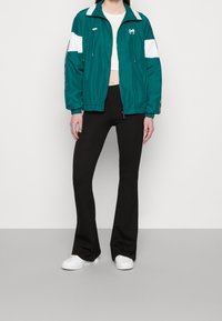 Niebieska kurtka zapinana na zamek z białymi akcentami, z kołnierzem typu stand-up i sznurkami, noszona z czarnymi spodniami dzwonami i białymi sneakersami.