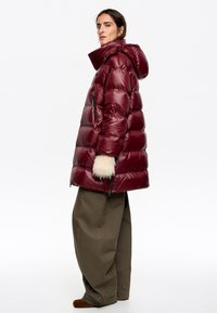 Bordowa pikowana kurtka puchowa z kapturem, z przeszywanymi panelami, bocznymi kieszeniami na zamek i futrzanymi mankietami. W zestawie z szerokimi khaki spodniami.