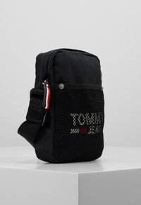 Borsa a spalla in nylon nero con tasca anteriore in rete, caratterizzata da un logo bianco e rosso. Spallaccio regolabile e scomparto principale con zip. Forma rettangolare.