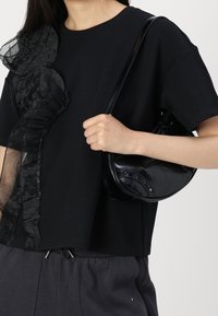 Černý krátký rukáv top s průsvitnými volánkovými detaily na levém rameni; modelka drží černou koženou kabelku s dekorativními kovovými prvky.