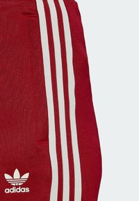 Rode Adidas trainingsbroek met drie witte strepen aan de zijkant, een gladde textuur en een klein wit logo op het onderste deel.