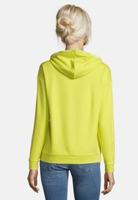 Kvinna med blont hår i en knut iförd en stark limegrön hoodie och blå jeans, sedd bakifrån mot en enkel bakgrund.