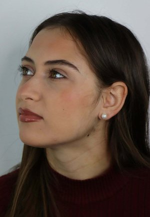 Giovane donna con capelli lunghi e scuri, che indossa orecchini a perno di perla e un dolcevita a coste color bordeaux, che guarda leggermente verso l'alto a sinistra.