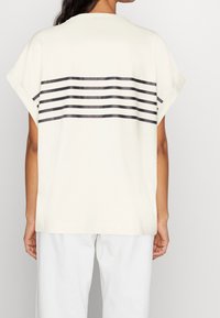 Kermanvärinen lyhythihainen T-paita pyöreällä pääntiellä ja harmaat vaakaraidat selässä. Pehmeä kangas, rento istuvuus.