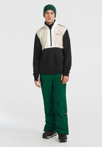 Heren gelaagde outfit: zwart-wit ritsjack met groene accenten, groene sneeuwbroek en zwarte sneeuwlaarzen. Eenvoudig ontwerp, functionele stof.