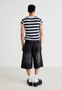 Persoană întoarsă, îmbrăcată cu un tricou cu dungi negre și albe, pantaloni scurți negri din denim lungi, șosete albe și pantofi cu talpă înaltă negri.