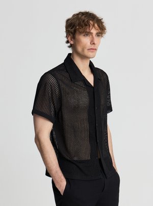 Uomo con capelli ricci che indossa una maglietta nera a maniche corte in rete e pantaloni neri, in piedi con le mani nelle tasche, guarda verso destra.