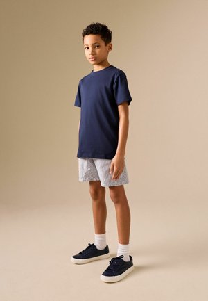 Junge steht und trägt ein marineblaues T-Shirt, hellgraue Shorts, weiße Socken und schwarze Turnschuhe vor einem einfarbigen beigen Hintergrund.