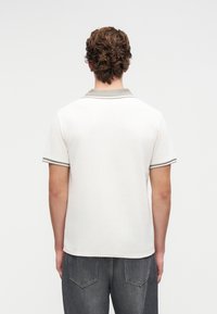 Mann in einem weißen Polohemd mit kurzen Ärmeln und grauen Besätzen am Kragen und an den Ärmeln, der mit dem Rücken zur Kamera steht und dunkle graue Jeans trägt.