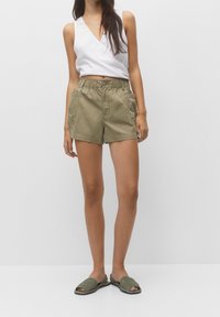 Shorts en coton vert olive avec une taille élastique, deux poches avant et un ourlet sur mesure. Portés avec un haut blanc sans manches et des sandales vertes.