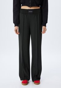 Zwarte, wijde broek met een elastische tailleband met het label "REPEAT." Gemaakt van glad materiaal, met naad aan de zijkanten, gecombineerd met rode schoenen.