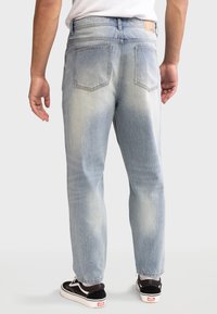 Jeans in denim azzurro chiaro con una vestibilità rilassata, dotati di due tasche posteriori e una toppa in pelle sulla vita, indossati con sneakers nere.