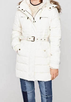 Person iført hvid quiltet vinterfrakke med bælte og pelsbeklædt hætte samt blue jeans med lapper, hænder delvist i lommerne.